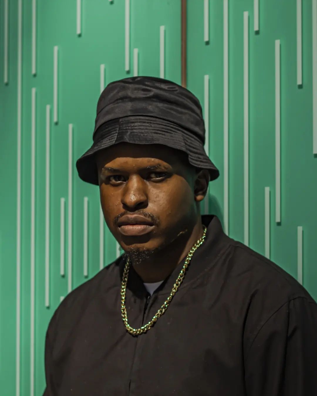 Kamo. G in a black bucket hat and jacket against a green background